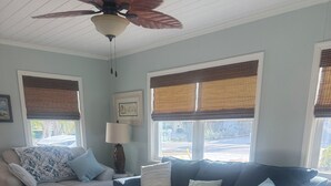 A living room with a beige sofa, a blue sofa, a patterned armchair, and a white ottoman. There are two windows with blinds, a ceiling fan, and a rug with a leaf pattern.