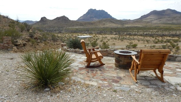 A patio with two wooden chairs and a stone fire pit overlooking a mountainous landscape. - Casa Tranquilla: Scenic Solar and Wind Powered Home (Alpine)