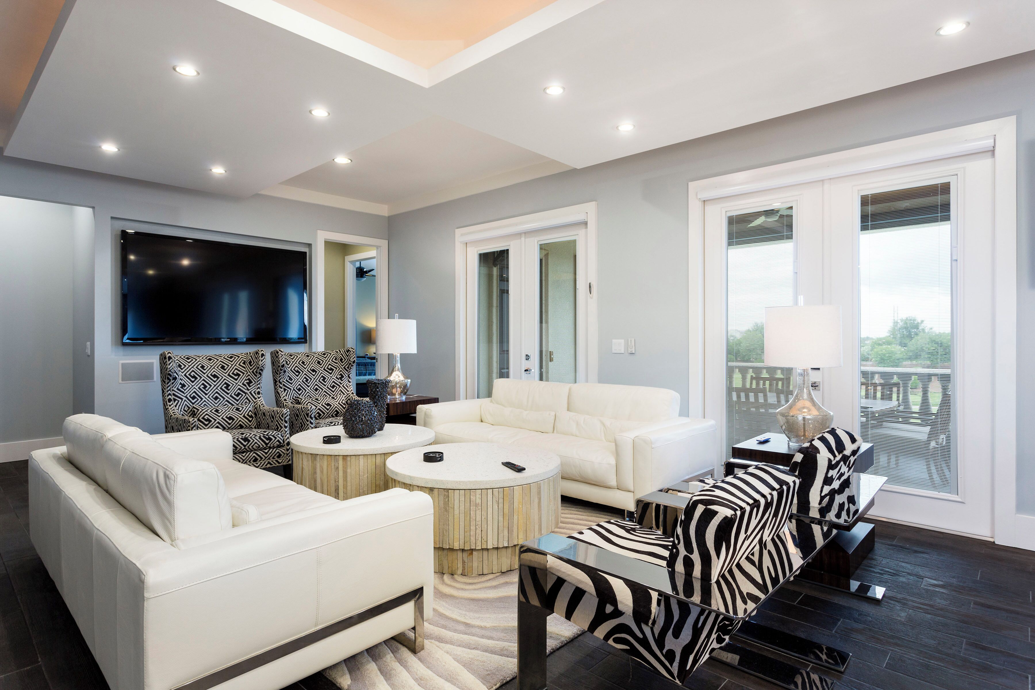 A modern living room with a flat-screen TV, white sofas, a zebra-patterned ottoman, and a glass coffee table.