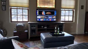 Een woonkamer met een flatscreen televisie op een houten standaard, twee ramen met zonwering, een zwarte leren bank, een kleine tafel met een mand en een decoratief dienblad met flessen.