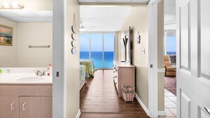A hotel room with a wooden floor, a large window with a view of the ocean, a bathroom with a sink and mirror, and a balcony with chairs. - Oceanfront 2-bedroom condo in Panama City Beach, sleeps 8, ideal for families. (Panama City Beach)