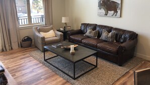 A living room with a brown leather sofa, a beige armchair, a coffee table, and a rug. There is a window with curtains, a lamp, and a framed picture on the wall. - Luxury 2 Bedroom, 2 Bath Condo - Perfect for a Fun Filled Family Get Away! (Mammoth Lakes)