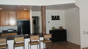A modern kitchen with wooden cabinets, a refrigerator, and a microwave. There is a dining area with wooden chairs and a black table. A bar area is visible with a 'BAR' sign on the wall. - Luxury 2 Bedroom, 2 Bath Condo - Perfect for a Fun Filled Family Get Away! (Mammoth Lakes)