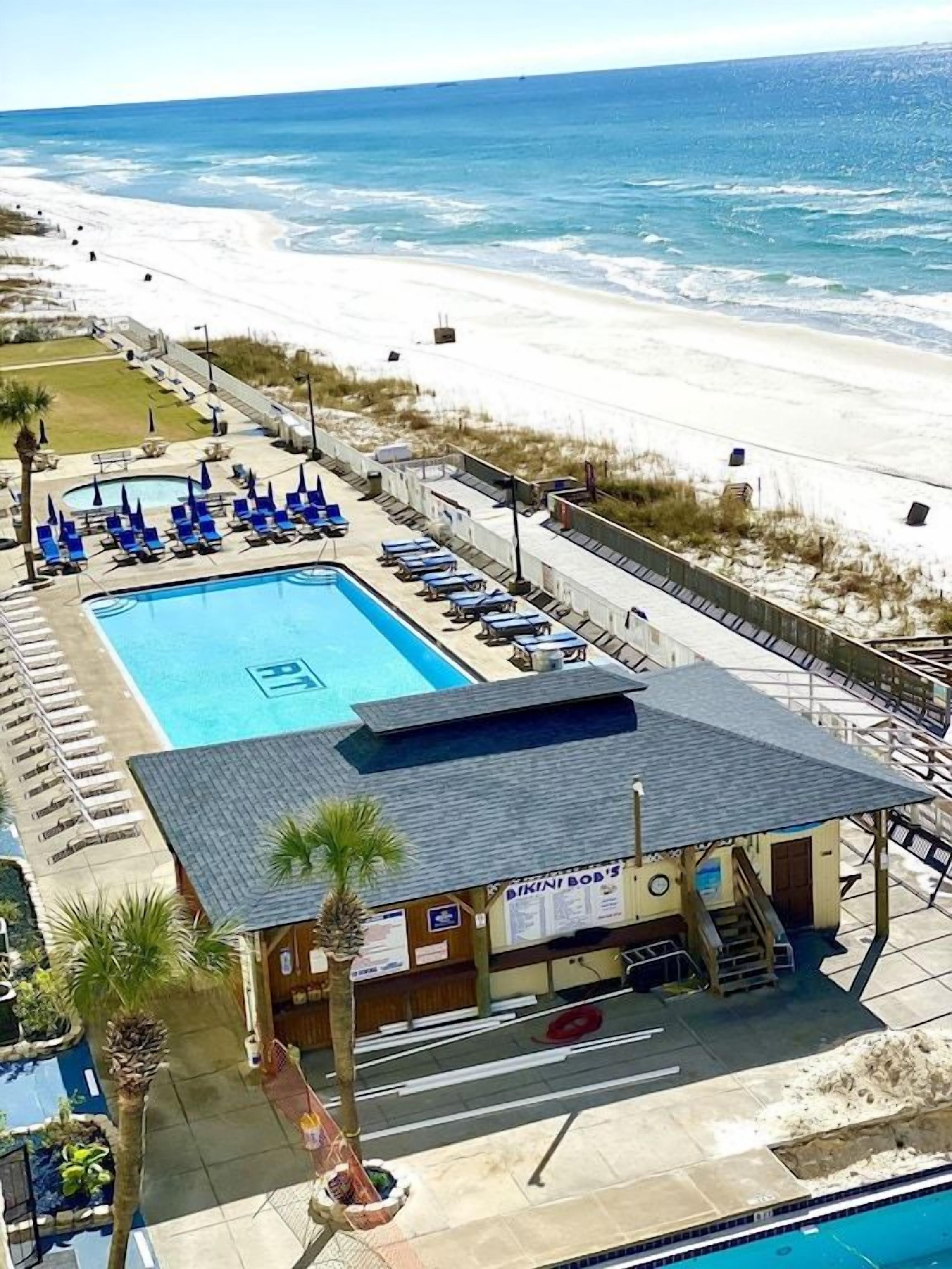 Zona de piscina con vista a la playa, sillas de descanso y un edificio con un cartel.