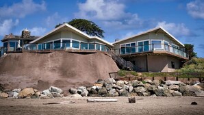 A modern coastal house with a large rock foundation, glass balconies, and a clear view of the ocean. - Captain's Quarters (Santa Cruz)