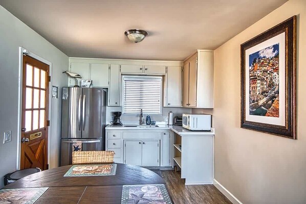 A compact kitchen with white cabinets, a stainless steel refrigerator, a microwave, and a window with blinds. - Captain's Quarters (Santa Cruz)