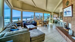 A spacious living area with a large window overlooking the ocean, a cozy seating area with a sofa and ottoman, a dining table with chairs, and a brick accent wall with a framed picture. - Captain's Quarters (Santa Cruz)