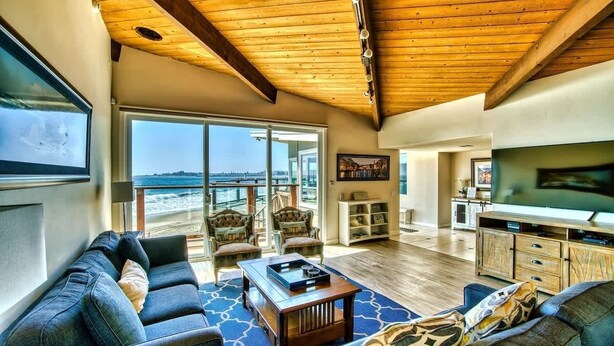 A spacious living room with a wooden ceiling, large windows, a television, and a view of the beach. - Captain's Quarters (Santa Cruz)