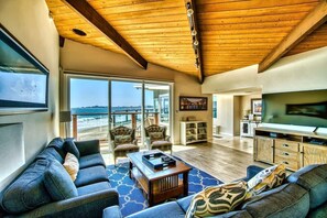 A spacious living room with a wooden ceiling, large windows, a television, and a view of the beach. - Captain's Quarters (Santa Cruz)