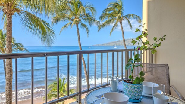 A balcony with a view of the ocean, palm trees, and a table set with white dishes and a potted plant.