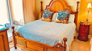 A wooden four-poster bed with a blue bedspread and patterned pillows, a wooden nightstand with a lamp, and a view of a window with curtains. - LUXURIOUS CONDO ON THE BEACH includes FREE BEACH CHAIRS. (Panama City Beach)