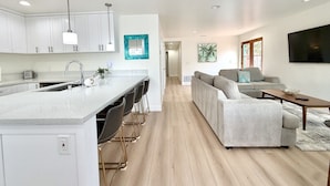 A modern kitchen with a central island, a dining area with bar stools, a living room with a sofa and a TV, and a hallway leading to another room. - JULY 2026 NEW REMODEL... Mermaid`s Cove, Nestled between Beach and Bay! (San Diego)