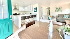 A modern kitchen with white cabinets, a central island with bar stools, and a dining area with a wooden table and chairs. - JULY 2026 NEW REMODEL... Mermaid`s Cove, Nestled between Beach and Bay! (San Diego)