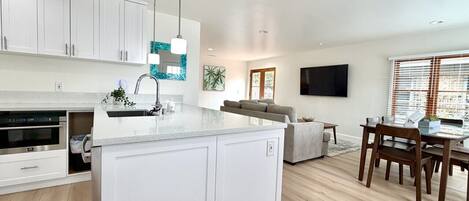 Una cocina moderna con muebles blancos, una isla central y una zona de comedor con sillas y mesa de madera.