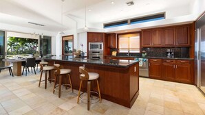 A modern kitchen with a large island, bar stools, wooden cabinets, stainless steel appliances, and a view of the outdoors.