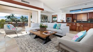 A modern living room with a sofa, armchairs, and a coffee table. There is a kitchen area with a bar and a large window overlooking the ocean.