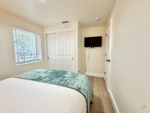 A bedroom with a bed, a window with blinds, a door, and a mounted TV.