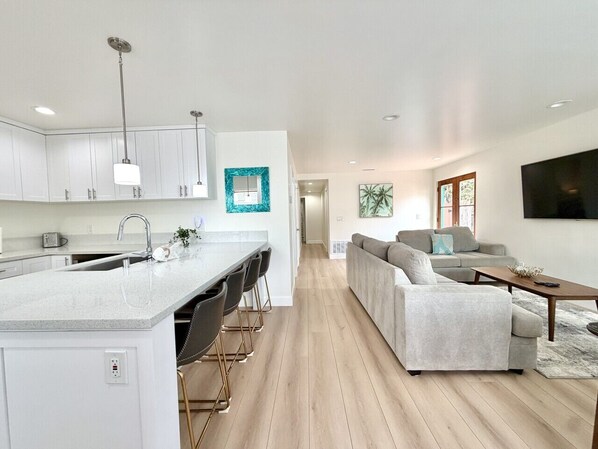A modern kitchen with a central island, a dining area with bar stools, a living room with a sofa and a TV, and a hallway leading to another room.