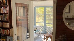 Una habitación acogedora con suelo de madera, una puerta blanca que da a un balcón, una pequeña mesa con una silla y una estantería llena de libros.