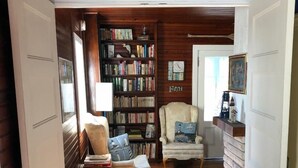 Una habitación acogedora con techo de madera, una estantería llena de libros, dos sillones y una pequeña mesa con una lámpara.
