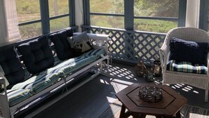 Un ambiente soleado con sofá, sillón de mimbre, una mesita y ventanas amplias con vistas a un paisaje natural.