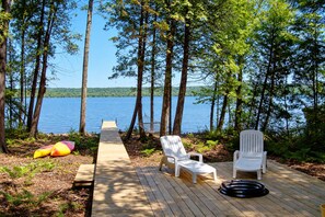 A wooden deck with a pathway leading to a lake, surrounded by trees, and featuring two white chairs and a hammock.