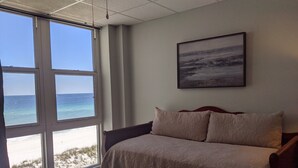 A bedroom with a large window overlooking a beach, a bed with a patterned bedspread, and a framed picture on the wall. - Directly on the Gulf, larger end unit,  both bedrooms have water view! (Pensacola Beach)