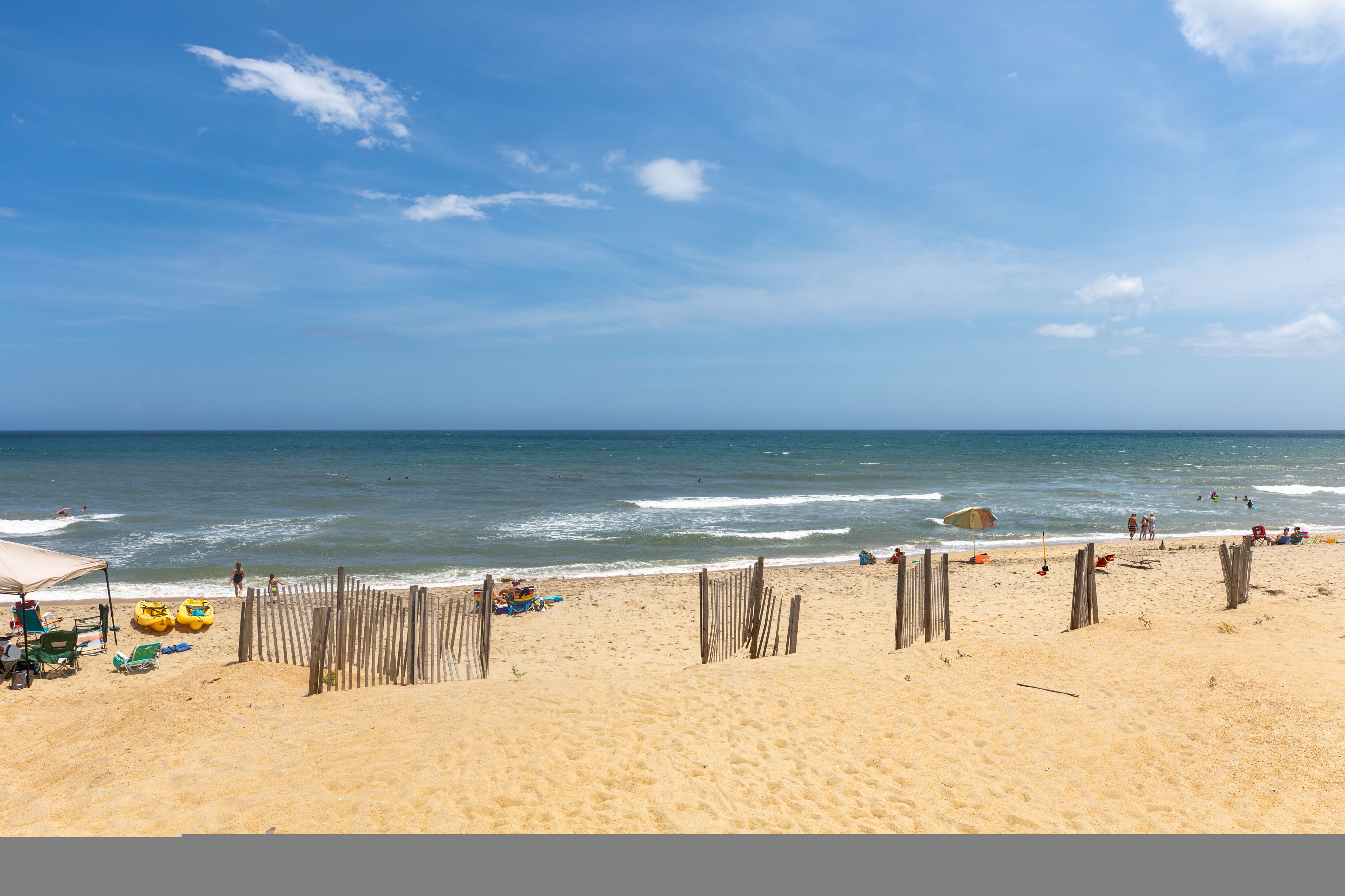 A sandy beach with a wooden fence, beach chairs, and a few people.