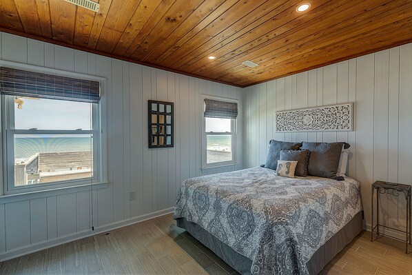 A bedroom with a large bed, wooden ceiling, and two windows with beach views.