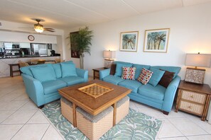 A living room with a blue sofa, a coffee table, and a kitchen in the background. - Experience comfort at Palm Tree Haven - Condo 504, a spacious 2BR retreat in South Padre Island. (South Padre Island)