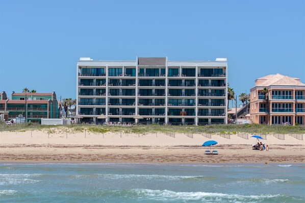 A multi-story hotel with balconies, located on a beach with a sandy shoreline and ocean view.