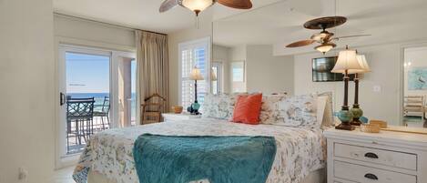A bedroom with a large bed, a ceiling fan, a dresser, and a view of the ocean through the balcony doors.