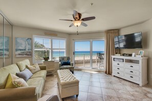 A living room with a sofa, ottoman, and a television on a stand. There is a ceiling fan, a window with a view of the beach, and a balcony with a chair. - Pets (dogs)Welcome *  Sea 4 Miles*  Sea 4 Yourself* Maravilla 2301 (Destin)