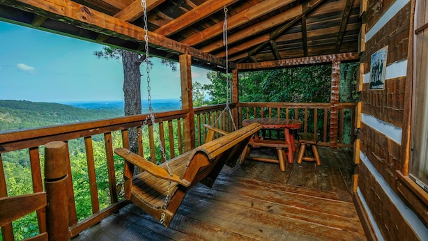 Une terrasse en bois avec un fauteuil suspendu, une table et un banc, d’où l’on peut admirer un paysage boisé.