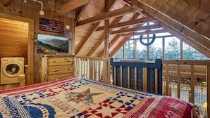 Une chambre confortable située sous les toits, avec un plafond en bois, un grand lit recouvert d’une couette aux couleurs vives, une télévision et une vue sur les arbres à travers de grandes fenêtres.