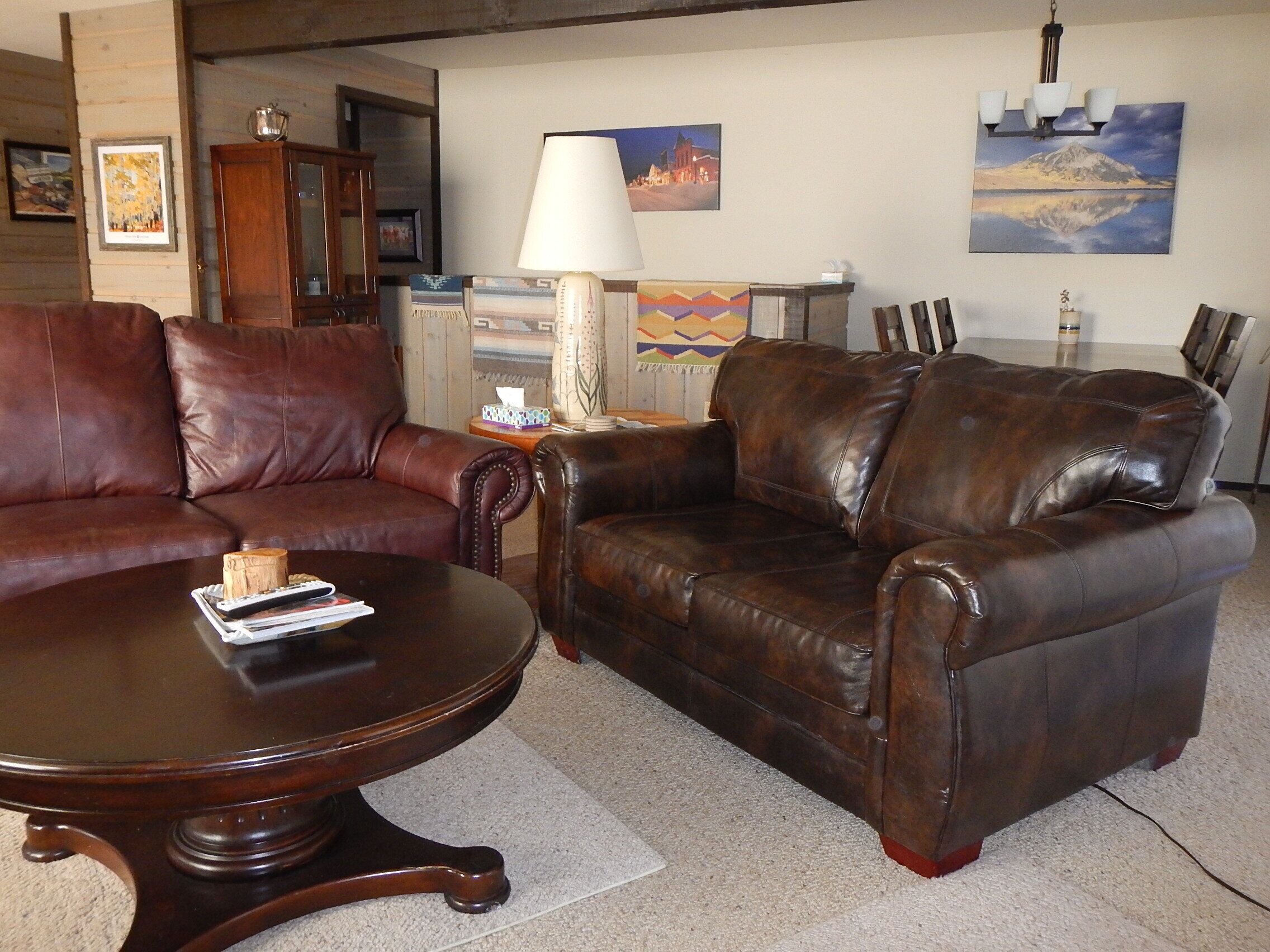 A living room with a brown leather sofa, a wooden coffee table, and a lamp. There are paintings on the wall and a wooden cabinet in the background.