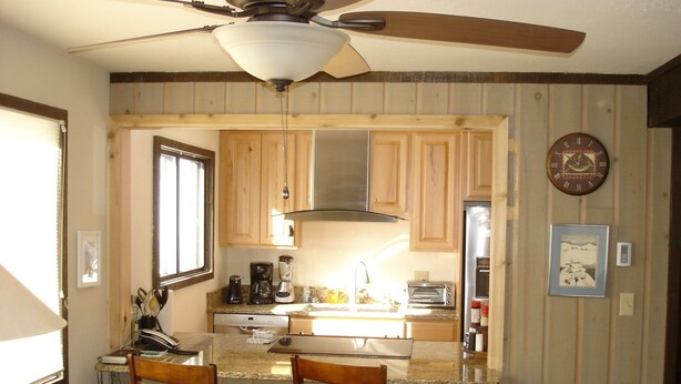 Une cuisine avec des meubles de rangement en bois, un ventilateur de plafond, un réfrigérateur, un grille-pain et une machine à café.