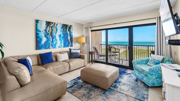 A modern living room with a beige sofa, a matching ottoman, and a colorful armchair. A large abstract painting adorns the wall, and a flat-screen TV is mounted on the wall. The room features a large sliding glass door leading to a balcony with a view of the ocean. - Experience the Island at our  Paradise Palm - Condo 304, a 2BR retreat in South Padre Island. (South Padre Island)