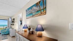 A living room with a wooden console table, two lamps, a painting of a beach scene, and a view of the ocean through a door. - Experience the Island at our  Paradise Palm - Condo 304, a 2BR retreat in South Padre Island. (South Padre Island)