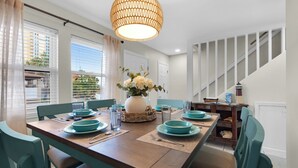 A dining area with a wooden table set for four, teal chairs, a white vase with flowers, and a hanging light fixture. - Beach Rendezvous-Beachside Complex with a Pool (Navarre)
