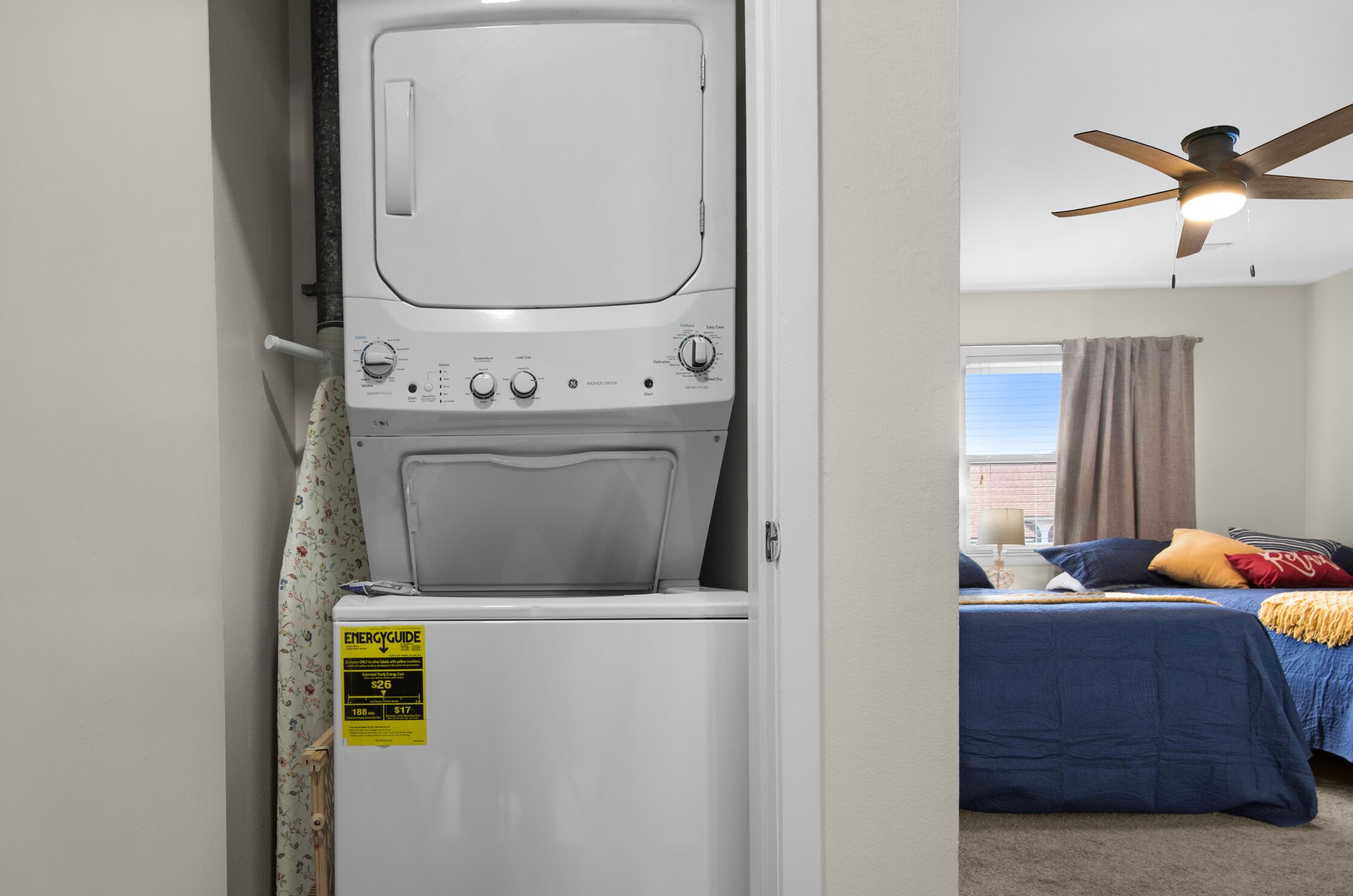 A laundry room with a front-loading washer and dryer, a ceiling fan, and a bedroom with a bed, pillows, and a nightstand.