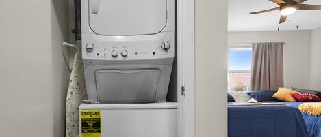 A laundry room with a front-loading washer and dryer, a ceiling fan, and a bedroom with a bed, pillows, and a nightstand.