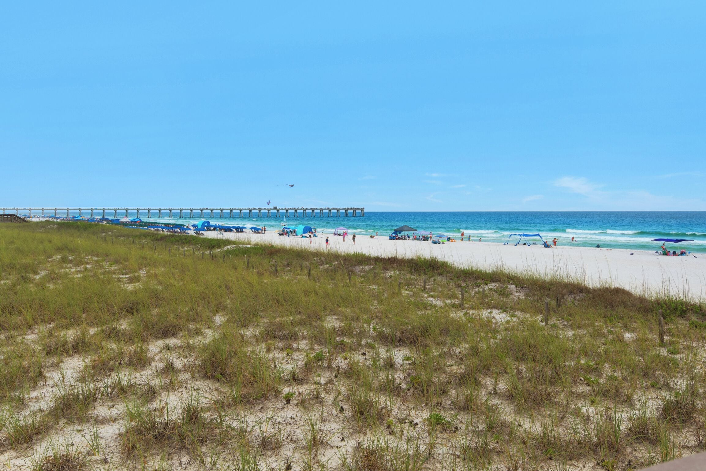 Une plage avec une jetée qui s’avance dans l’océan, des dunes de sable et un ciel bleu dégagé.