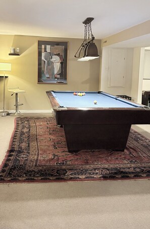 A pool table with a hanging light fixture, a framed abstract painting, and a patterned rug.