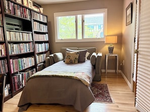 A bedroom with a large bookshelf, a bed with a patterned bedspread, a window with a view of buildings, and a small table with a lamp.