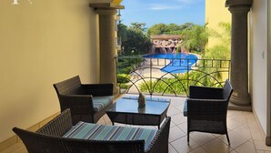 A balcony with wicker furniture, a view of a pool, and a tropical landscape.