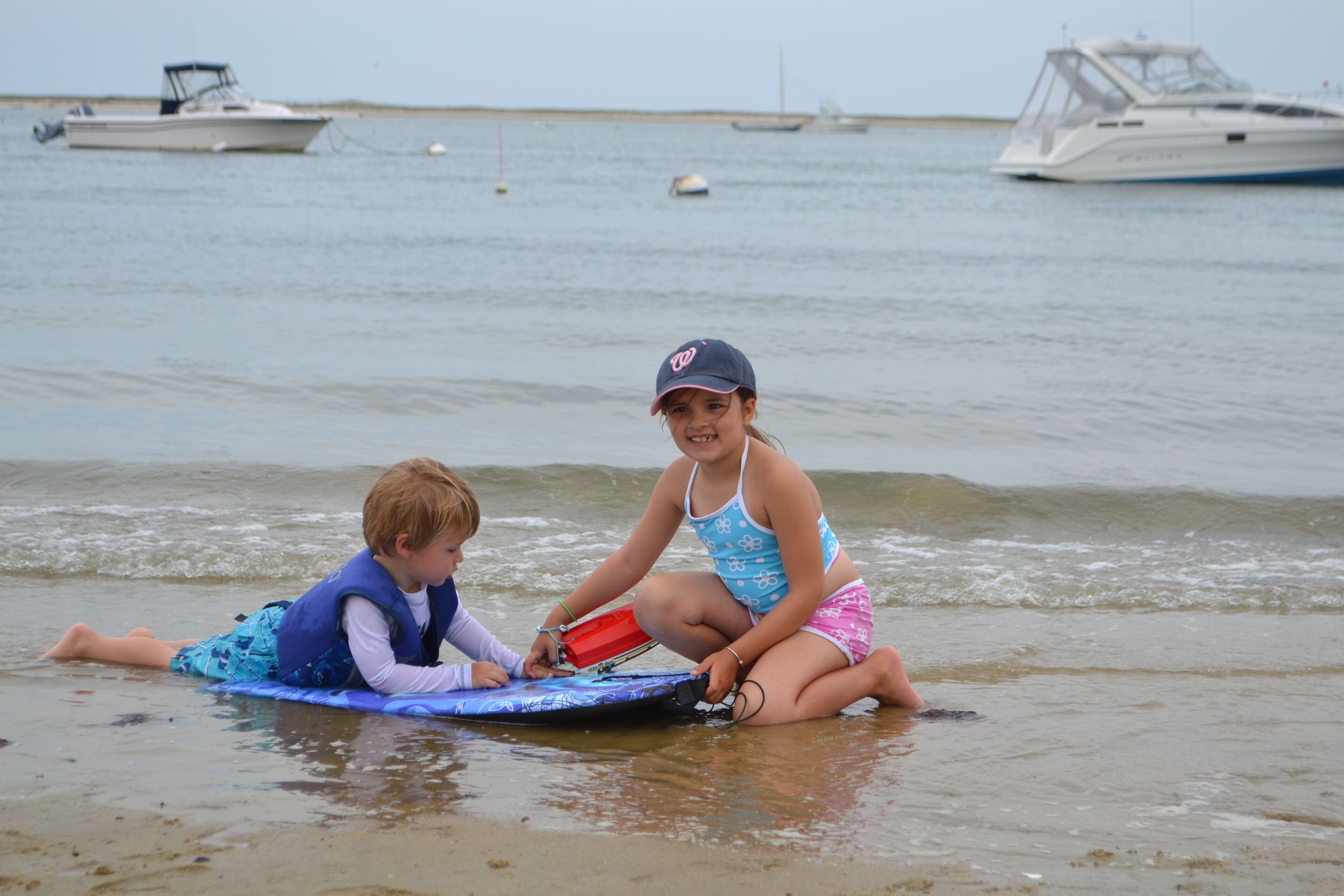 Two children on a surfboard in shallow water with boats in the background.