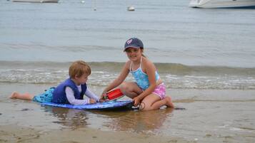 Two children on a surfboard in shallow water with boats in the background.