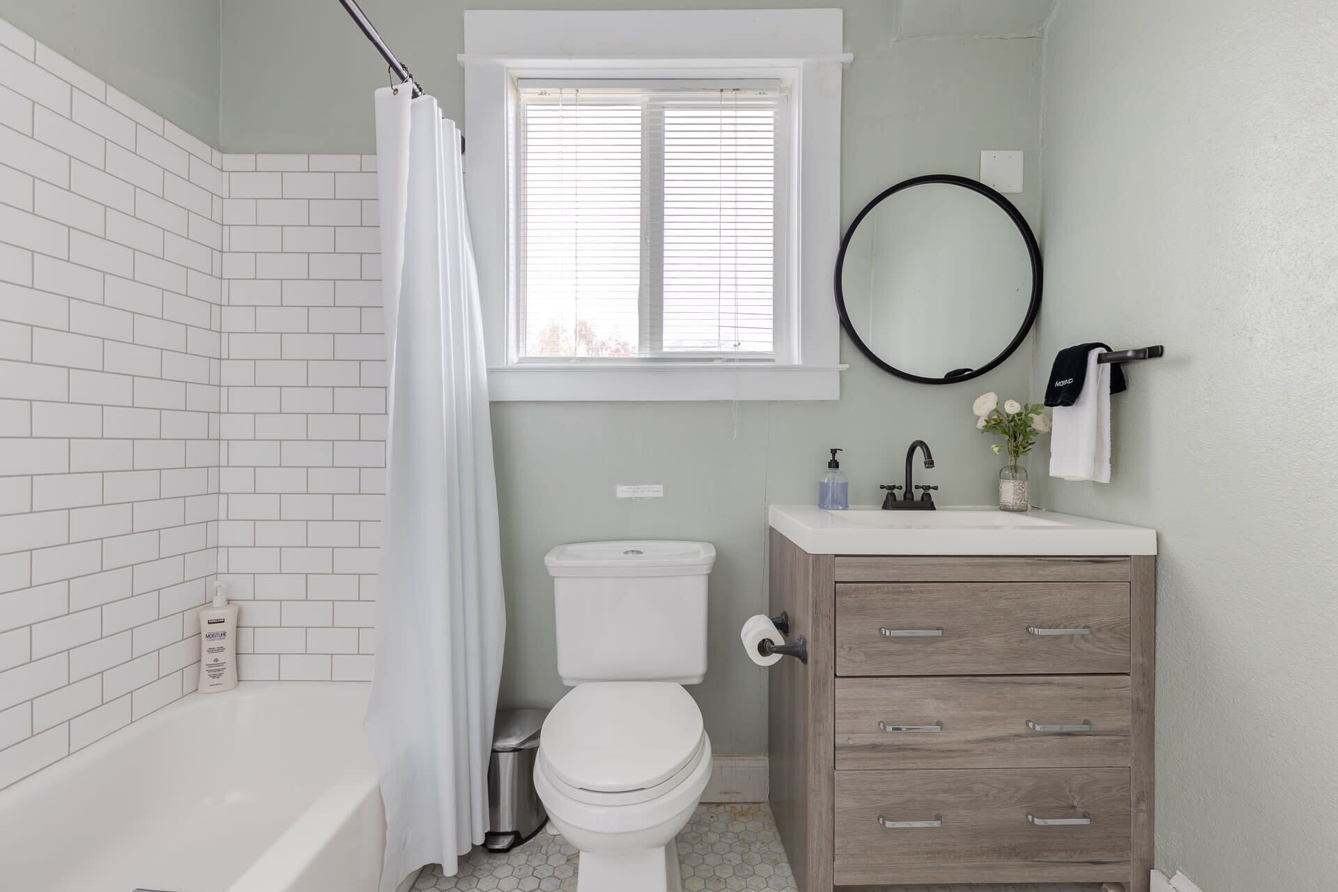 A bathroom with a white bathtub, a toilet, a vanity with drawers, a window with blinds, and a mirror with a towel hanging on it.