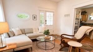 A living room with a sofa, a coffee table, a chair, a lamp, and a window with curtains. - Historic Craftsman, Walk to Duke (Durham)